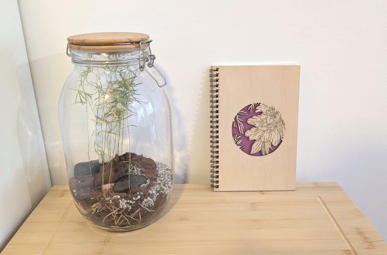 Puede incluir: Un gran terrario de vidrio con tapa de madera y cierre met&aacute;lico, que contiene tierra, rocas y vegetaci&oacute;n. Un cuaderno de espiral de madera con un dise&ntilde;o floral est&aacute; junto al terrario. El cuaderno tiene un c&iacute;rculo morado con un dise&ntilde;o floral.