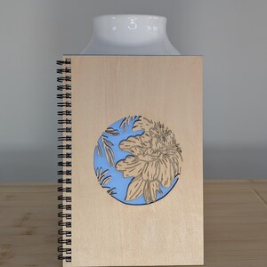Puede incluir: Un cuaderno de espiral de madera con un dise&ntilde;o floral cortado con l&aacute;ser. La cubierta es de color marr&oacute;n claro con un c&iacute;rculo azul con una flor y hojas. El cuaderno est&aacute; sobre una superficie de madera.