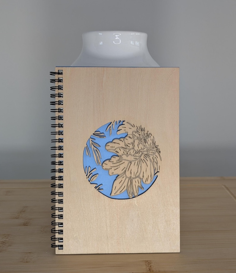 Puede incluir: Un cuaderno de espiral de madera con un dise&ntilde;o floral cortado con l&aacute;ser. La cubierta es de color marr&oacute;n claro con un c&iacute;rculo azul con una flor y hojas. El cuaderno est&aacute; sobre una superficie de madera.