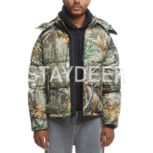 Puede incluir: Chaqueta acolchada con capucha y estampado de camuflaje en tonos verdes, marrones y beige. La chaqueta tiene una cremallera negra y la palabra "STAYDEER" impresa en la parte delantera. Se lleva sobre una sudadera con capucha negra.