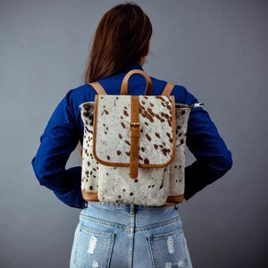 Puede incluir: Una mochila con un estampado de piel de vaca en blanco y marrón. La bolsa tiene correas de cuero marrón, un asa superior y una solapa frontal con cierre de hebilla. La mochila se lleva sobre los hombros.