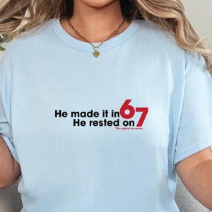 May include: Light blue t-shirt with the text "He made it in 67, He rested on". The text is in black and red. The shirt is made of a soft material and is suitable for casual wear.