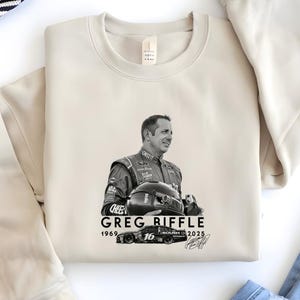 May include: A cream-coloured sweatshirt featuring a black and white image of Greg Biffle, a race car driver, holding a helmet. The text "GREG RIFFLE 1969-2025" is printed below the image, along with a race car graphic and signature.