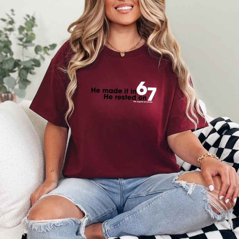 May include: A maroon t-shirt with the text "He made it in 67 He rested on" in black. The shirt is worn with blue jeans. The text is in a simple sans-serif font.