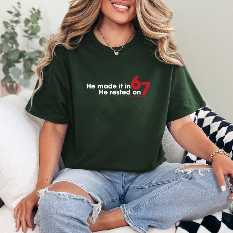 May include: Dark green t-shirt with the text "He made it in 67 He rested on" in white and red. The shirt is a crew neck and is made of a soft material. The model is wearing blue jeans.