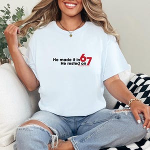 May include: A light blue t-shirt with the text "He made it in 67, He rested on the original six acres" in black and red. The shirt is made of a soft material.