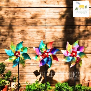 Puede incluir: Tres molinillos de viento coloridos con detalles brillantes se muestran frente a un fondo de madera. Cada molinillo tiene una combinación de colores diferente, con centros de flores rojas y estacas de metal negro. Un adorno decorativo para el jardín.