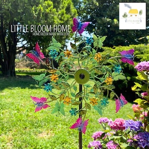 Puede incluir: Un molinillo de jardín decorativo de metal con mariposas, flores y hojas de colores. El molinillo tiene un marco verde y está situado en una zona exterior cubierta de hierba. El texto "LITTLE BLOOM HOME HOME DECOR MADE WITH LOVE" es visible.