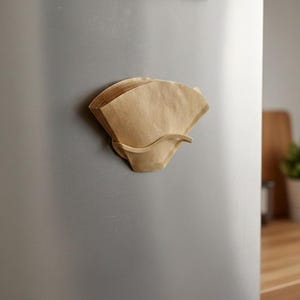 Può includere: Un portafiltro per caffè magnetico in legno chiaro, che contiene una pila di filtri di carta marrone. Il supporto è attaccato a una porta del frigorifero in acciaio inossidabile. Un disegno per bambini è attaccato sopra.