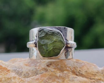 May include: A sterling silver ring featuring a rough-cut, olive-green gemstone. The ring has a wide band with a textured finish and a bezel setting that highlights the natural shape of the stone. The ring is displayed on a light brown surface.