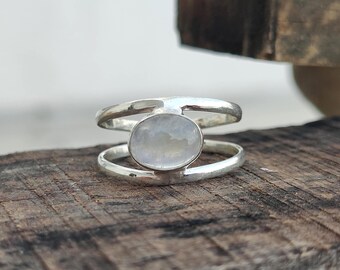 May include: A silver ring with a large, oval, iridescent gemstone. The ring has a double-band design, with the gemstone set between the two bands. The ring is displayed on a wooden surface. The text "925 Silver Legacy" is visible.