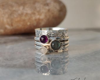 May include: A silver spinner ring with multiple bands and floral detailing. The ring features three gemstones: a purple amethyst, a gray labradorite, and a white moonstone. The ring is displayed on a reflective surface.