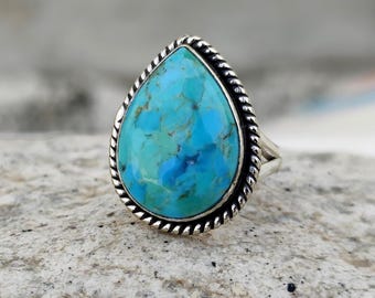 May include: A sterling silver ring featuring a large, teardrop-shaped turquoise stone. The stone is a vibrant mix of blue and green hues, set within a twisted rope-like border. The ring is displayed on a textured, light-colored surface.