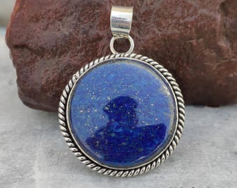 May include: A round, blue lapis lazuli pendant with a silver-colored rope-like border. The pendant has a silver bail for a chain. The stone has flecks of gold and is set against a natural background.