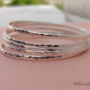 May include: A stack of four silver bangles with a hammered texture. The bracelets are stacked on a pink surface, with the brand name "925 Silver Legacy" in the bottom right corner. The bangles are a simple, elegant design.