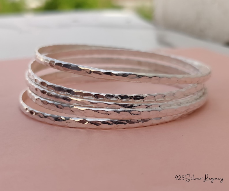 May include: A stack of four silver bangles with a hammered texture. The bracelets are stacked on a pink surface, with the brand name "925 Silver Legacy" in the bottom right corner. The bangles are a simple, elegant design.
