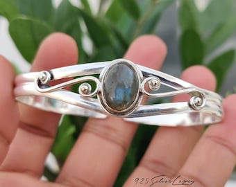 May include: A sterling silver cuff bracelet featuring a central oval-shaped labradorite gemstone with blue iridescence. The band is crafted with polished silver wire, showcasing decorative swirling metalwork and small silver spheres on each side.