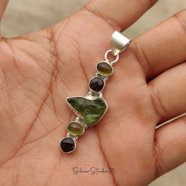 May include: A silver pendant featuring a rough-cut, olive-green gemstone at the center, flanked by smaller, round, dark red and olive-green stones. The pendant has a loop at the top for a chain. The text "Silver Studio 25" is at the bottom.