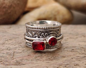 May include: A silver spinner ring with two red gemstones. One gemstone is round, and the other is square. The ring has an engraved floral design and the text "Silver Studio 25".