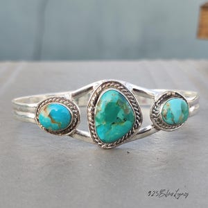 May include: A silver bracelet with three turquoise stones. The central stone is a larger, irregular shape, with two smaller, round stones on either side. The turquoise is a mottled blue-green colour. The bracelet has a braided design around the stones.
