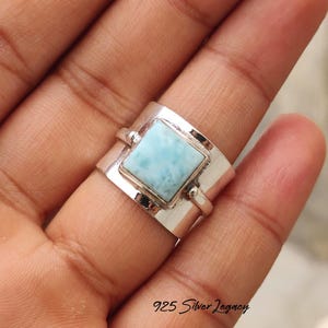 May include: A wide silver ring featuring a square, light blue gemstone. The ring has a modern design with a rectangular band and a bezel-set stone. The text "925 Silver Legacy" is visible at the bottom.