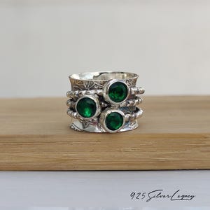 May include: A silver ring with three green gemstones. The ring has a detailed floral design and the text "925 Silver Legacy" is visible at the bottom. The gemstones are round and set in a silver band.
