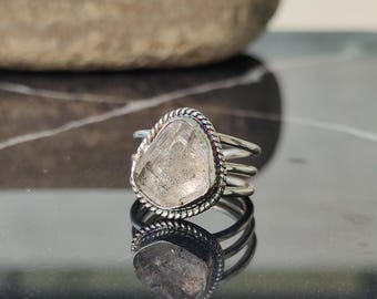 May include: A silver ring with a large, irregularly shaped, clear gemstone. The gemstone is set in a twisted rope-like bezel. The ring has a triple-band design. The ring is reflected in the glossy surface below. The text "925 Silver Legacy" is at the bottom.