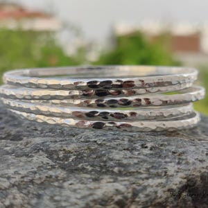 May include: A stack of five hammered silver bangles. The bangles have a textured surface and a shiny, reflective finish. The jewelry is displayed on a gray stone surface with a blurred green background.