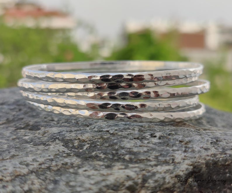 May include: A stack of five hammered silver bangles. The bangles have a textured surface and a shiny, reflective finish. The jewelry is displayed on a gray stone surface with a blurred green background.