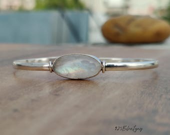 May include: A silver bangle bracelet with an oval, iridescent moonstone center. The bracelet has a simple, elegant design, perfect for everyday wear. The moonstone displays a play of colors, reflecting light. The bracelet is displayed on a wooden surface.
