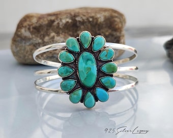 May include: A sterling silver cuff bracelet featuring a large central oval turquoise stone surrounded by twelve smaller teardrop-shaped turquoise stones in a cluster design. The metal band consists of three parallel silver wires. Text reads 925 Silver Legacy.