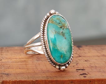 May include: A silver ring featuring a large, oval-shaped turquoise stone with brown matrix veining. The stone is set in a twisted rope border with small silver bead accents at the top and bottom. The band consists of three split silver shanks.