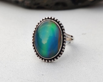 May include: A silver ring with an oval-shaped, color-changing stone. The stone transitions from green to blue, with hints of purple and yellow. The ring has a beaded border around the stone.