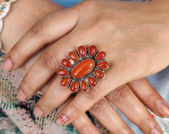 May include: A silver ring with a large, oval, orange-red stone at the center, surrounded by smaller, teardrop-shaped stones of the same color. The ring is on a hand, with the text "925 Silver Legacy" visible in the background.