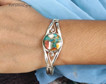 May include: A silver cuff bracelet with a central, round turquoise and multi-colored stone. The bracelet has a decorative, open design with swirling details. The text "925 Silver Legacy" is visible in the upper left corner.