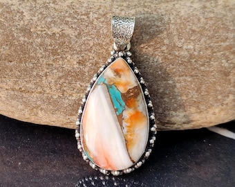 May include: A teardrop-shaped pendant with a silver bail. The pendant features a colorful stone with white, orange, and turquoise hues, set in a silver bezel with a beaded edge. The text "Silver Legacy" is at the bottom.