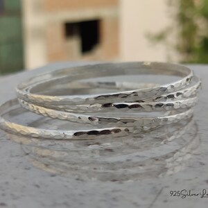 May include: A stack of four silver bangles with a hammered texture. The bangles are round and reflect light, creating highlights and shadows. The image is taken in natural light, with a blurred background.