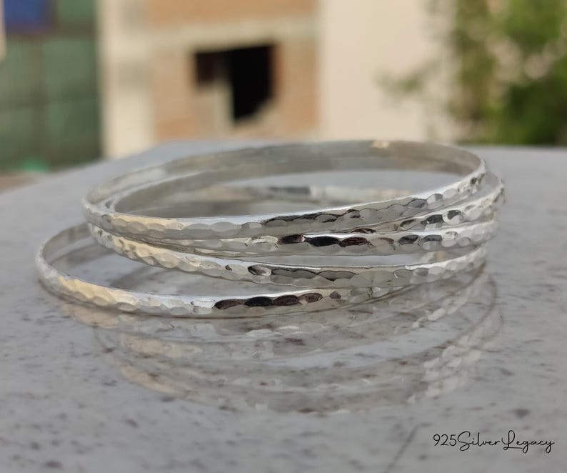 May include: A stack of four silver bangles with a hammered texture. The bangles are round and reflect light, creating highlights and shadows. The image is taken in natural light, with a blurred background.