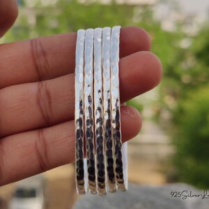 May include: A set of six hammered silver bangles. The bangles are held in a hand, showcasing their textured surface and reflective quality. The background is blurred, with green foliage visible.