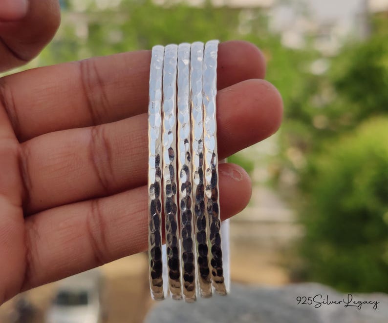 May include: A set of six hammered silver bangles. The bangles are held in a hand, showcasing their textured surface and reflective quality. The background is blurred, with green foliage visible.