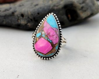 May include: A sterling silver ring with a teardrop-shaped stone. The stone features a vibrant mix of colors, including bright pink, turquoise, and bronze. The ring has a beaded border and a polished finish. The text "925 Silver Legacy" is visible.