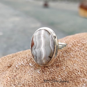 May include: A silver ring with an oval-shaped stone. The stone features a pattern of gray and white bands with a touch of red. The ring is set on a textured surface. The text "925 Silver Legacy" is visible.