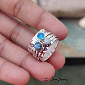 May include: A silver ring featuring three gemstones in blue, grey, and white. The ring has a floral design on the band. The text "925 Silver Legacy" is visible at the bottom.