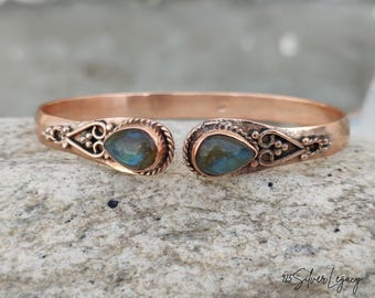 May include: A copper-colored cuff bracelet with ornate detailing and two teardrop-shaped, iridescent gemstones. The bracelet features decorative swirls and a twisted rope design near the gemstone settings. The jewelry is displayed on a textured, gray surface.