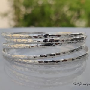 May include: A stack of silver bangles with a hammered texture. The bangles are stacked on a reflective surface, with a blurred green background. The jewelry is simple and elegant.