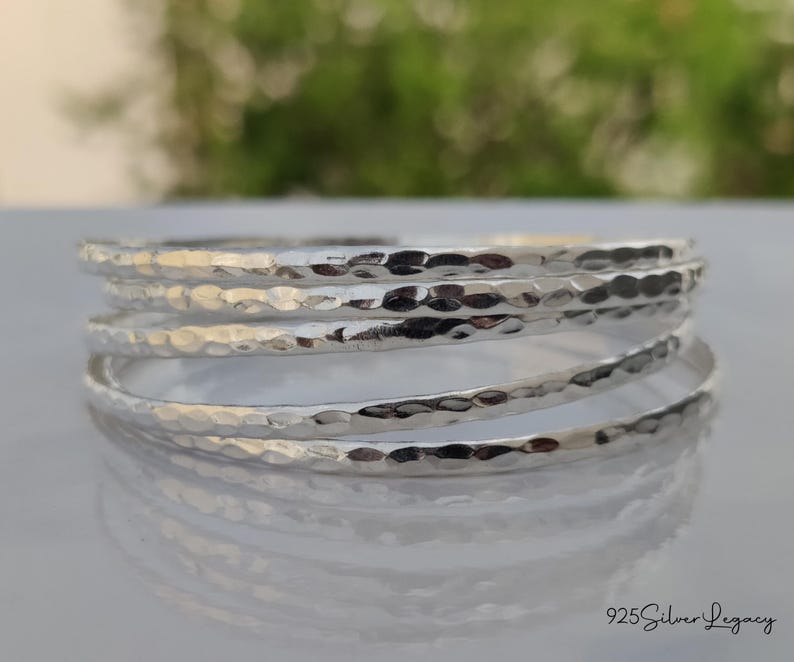May include: A stack of silver bangles with a hammered texture. The bangles are stacked on a reflective surface, with a blurred green background. The jewelry is simple and elegant.