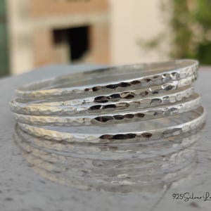 May include: A stack of five silver bangles with a hammered texture. The bangles are round and reflect light, creating a bright and shiny appearance. The jewelry is displayed on a light-colored surface.