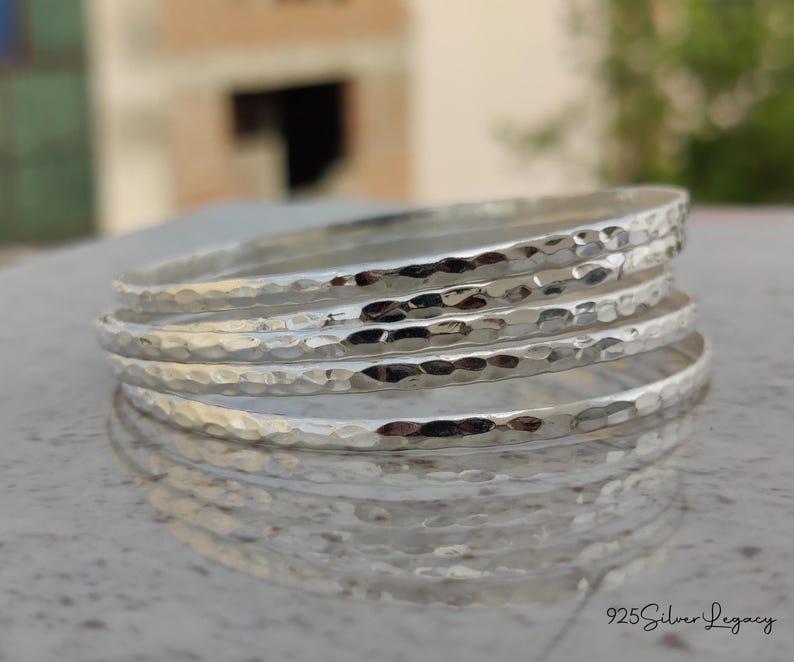 May include: A stack of five silver bangles with a hammered texture. The bangles are round and reflect light, creating a bright and shiny appearance. The jewelry is displayed on a light-colored surface.