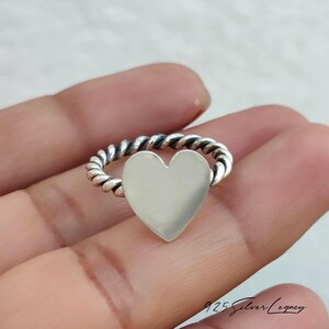 May include: A silver ring featuring a heart-shaped centerpiece. The band has a twisted rope design. The ring is held in a hand, with the focus on the jewelry. The ring is a piece of fashion jewelry.