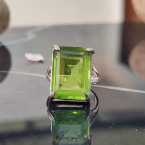 May include: A silver ring featuring a large, rectangular, light green gemstone. The gemstone is the focal point, set in a silver band with a simple, elegant design. The ring is displayed on a reflective surface.